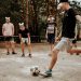 Group of teenagers wearing masks playing a soccer game in a sandy outdoor setting.